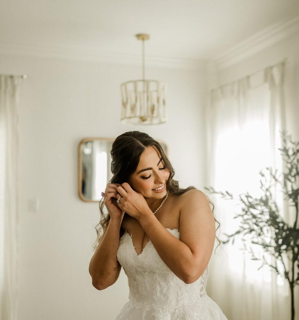 A bride is putting on her earrings in a room with white walls and minimalist but elegant decorations.