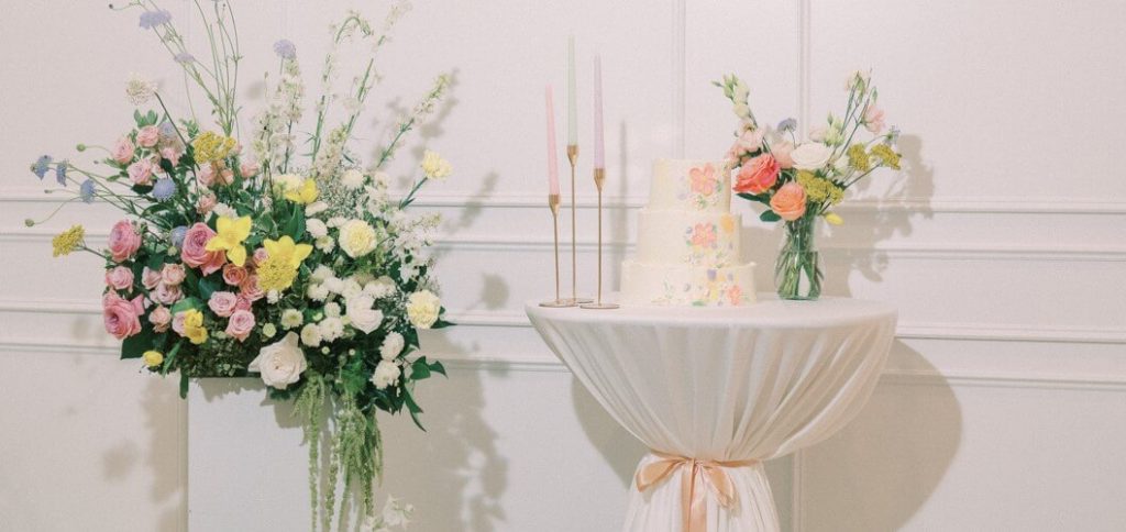 A cake sitting on a decorated cocktail table and a pillar of flower arrangements.