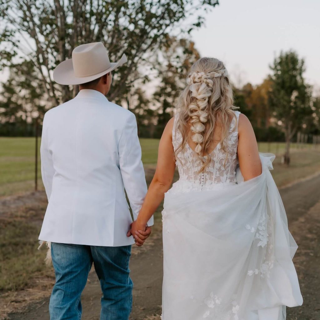 A groom and a bride walking at the ranch while holding each other's hands.