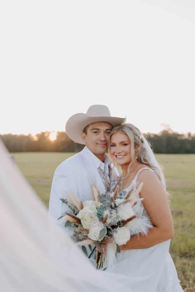 A lovely couple on their wedding day wearing matching white dress and suit, with the bride holding her bouquet and the groom wearing his cowboy hat.