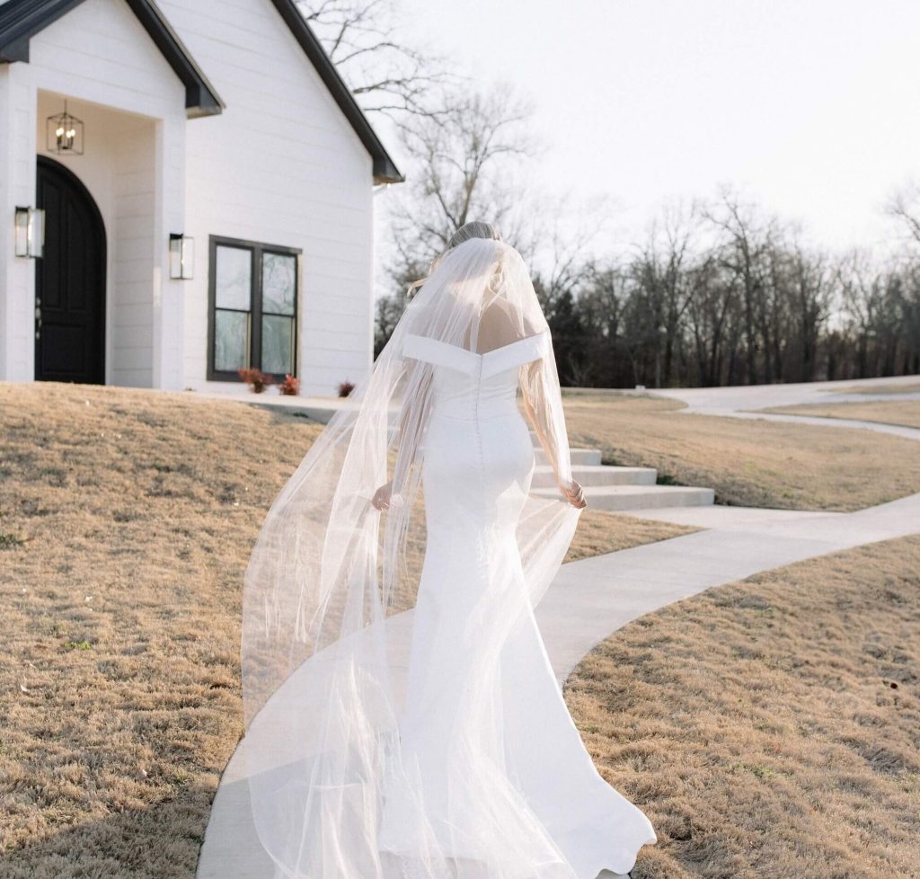 A bride wearing her off-the-shoulder satin mermaid wedding dress and veil walks on a path towards the church.