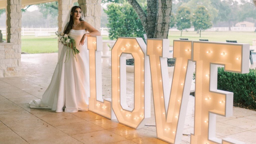 A bride is leaning on a big "LOVE" letter standee in her reception venue while holding her bouquet.