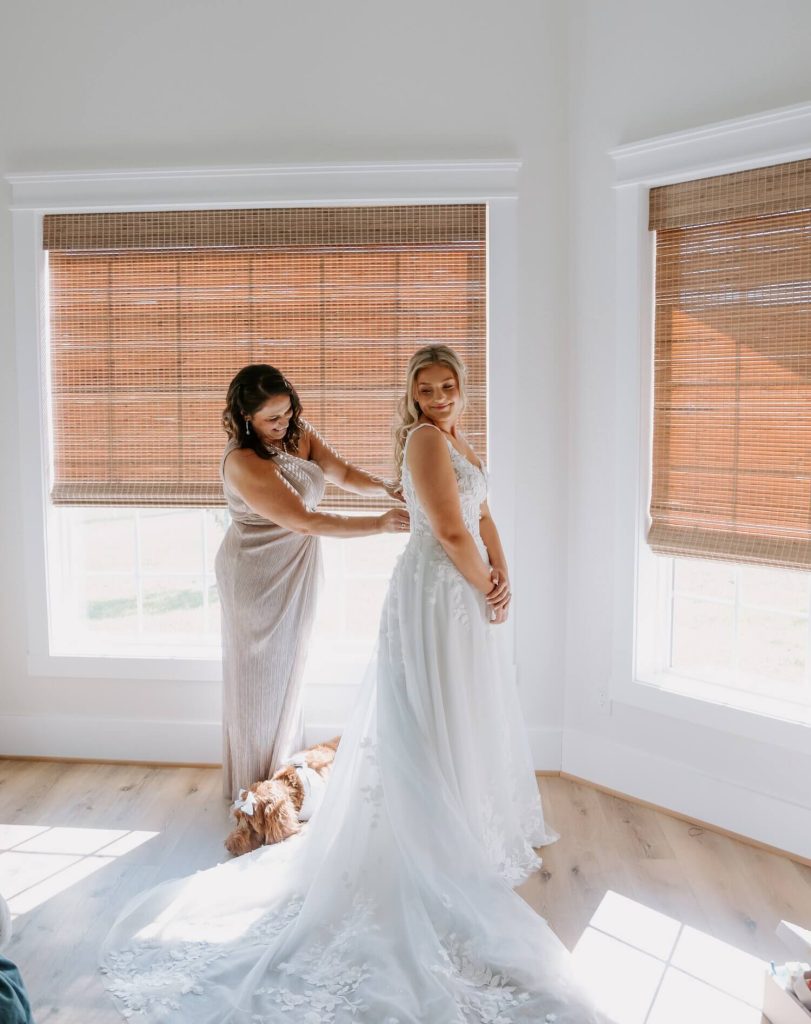 A mom helping her daughter get into her wedding dress in a white room with big windows and roll-up blinds.