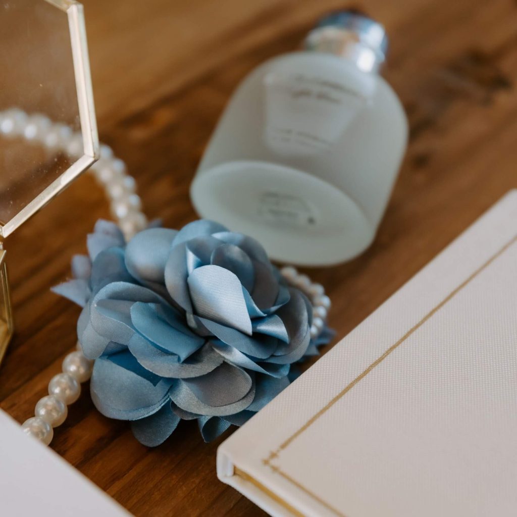 A blue fabric flower with a guest book, a perfume, a jewelry box, and a pearl necklace are laid on a wooden table.