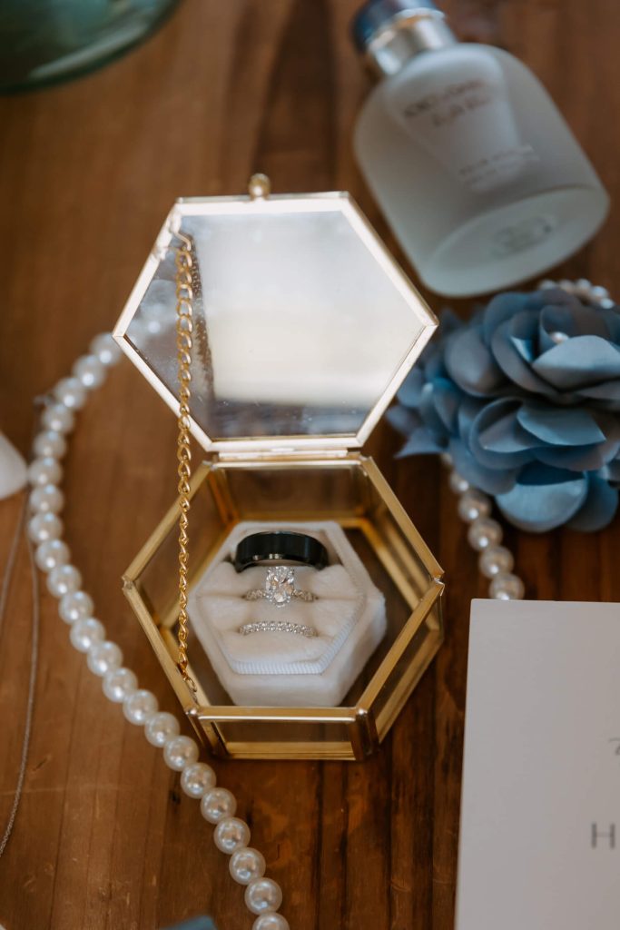 Wedding and engagement bands are placed in a hexagonal clear jewelry box with a pearl necklace, a perfume, and a blue flower around it.