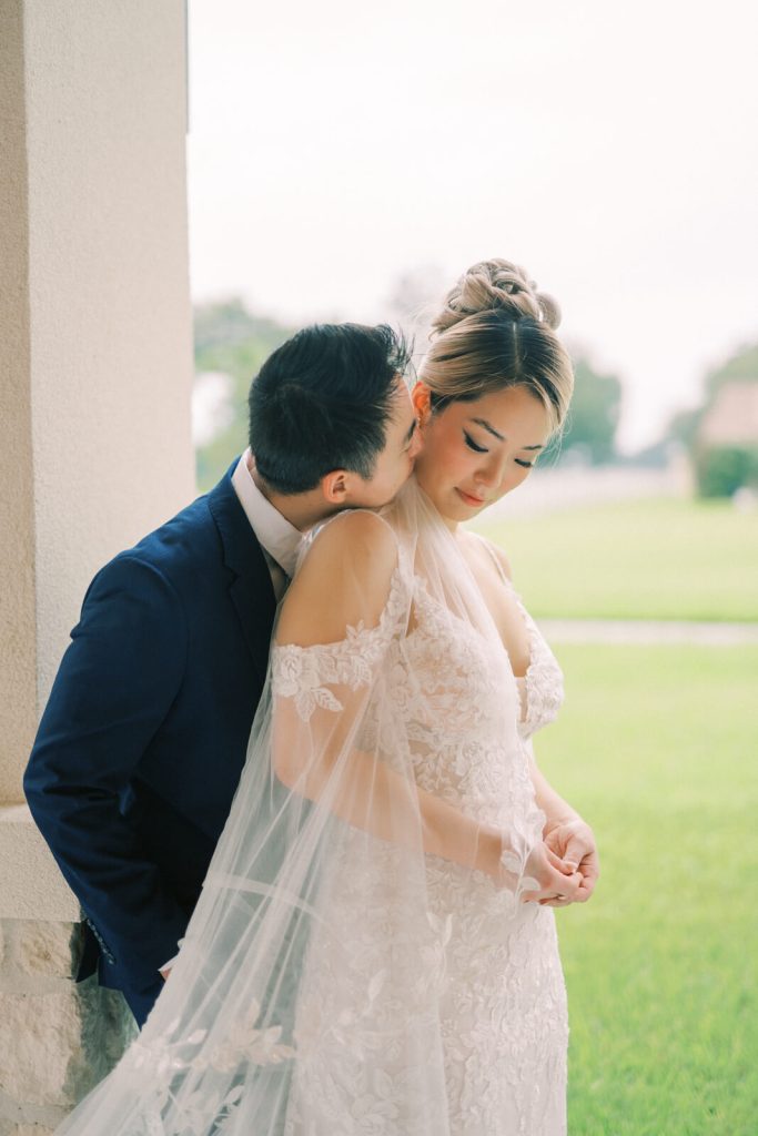 A portrait of a groom kissing his bride from behind while softly leaning on a concrete pillar.