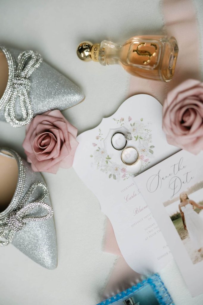 A pair of silver shoes, Jimmy Choo perfume, a wedding invitation, wedding bands, and two flowers are all arranged on a white surface.