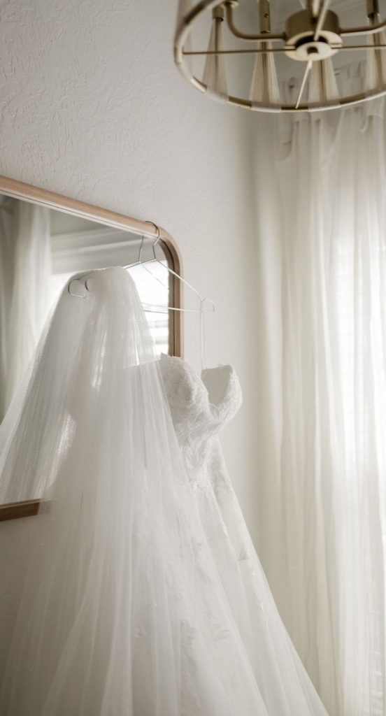 A white wedding dress with a sweetheart neckline is hung on a mirror frame beside the window.