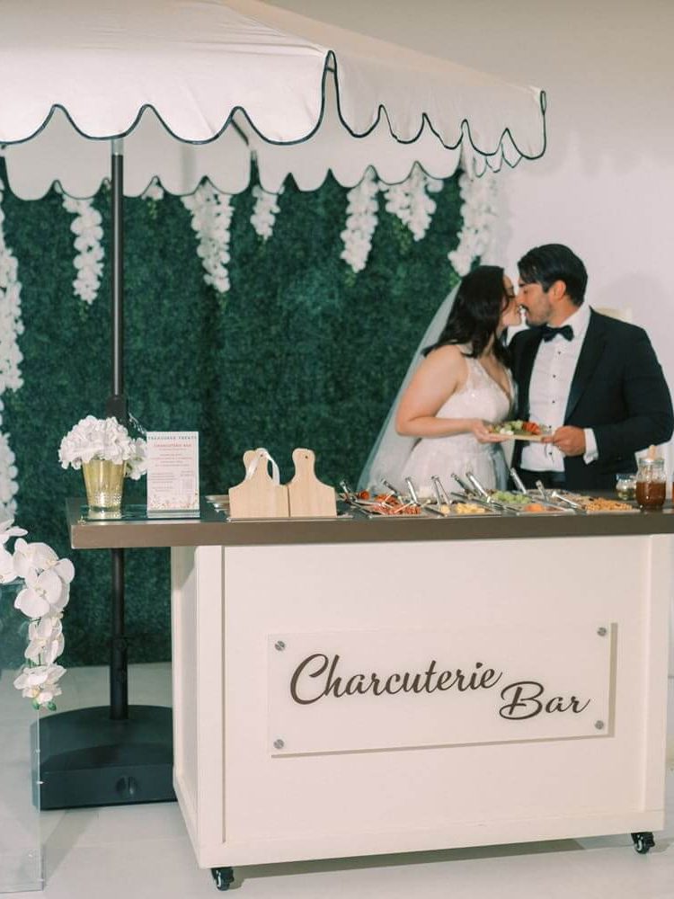 The bride and groom are leaning towards each other for a kiss by the charcuterie bar.