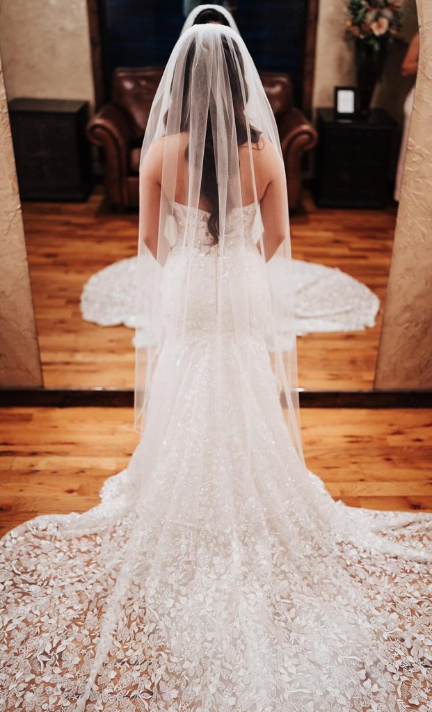 A bride wearing a sparkly lace mermaid wedding dress and a veil, looking at herself in the mirror.