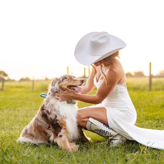 A bride in a white dress and wide-brimmed hat kneels in a grassy field, smiling as she pets a dog.