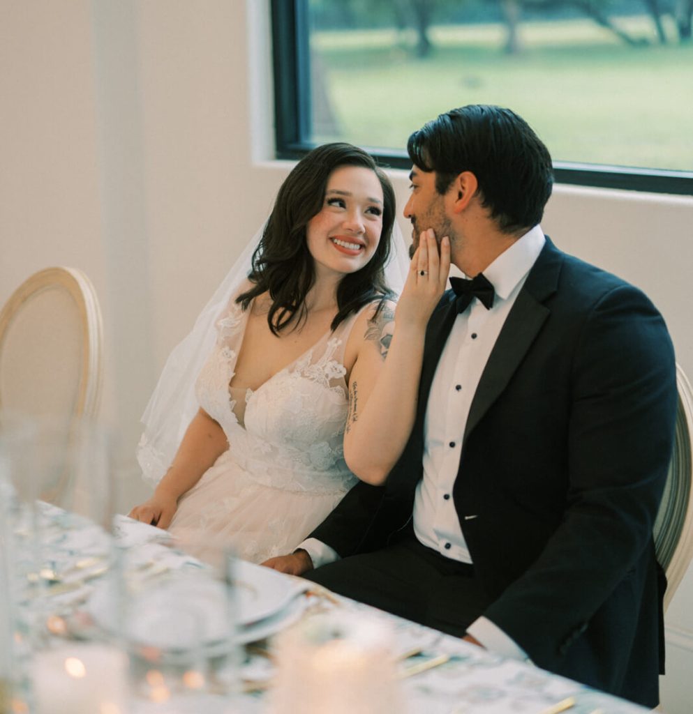The groom and bride are sitting on their sponsor's table with the bride's hands touching the groom's face.