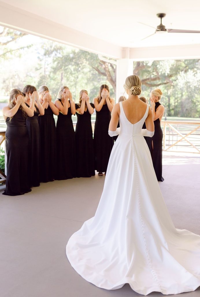 Bride in a long white gown walking toward her bridesmaids dressed in black for a first look moment.