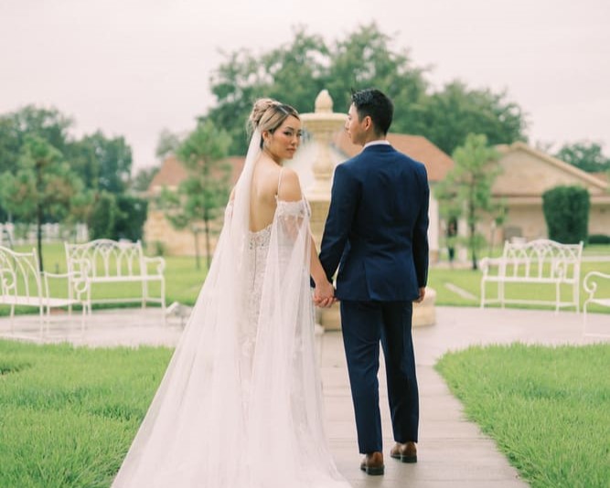 The groom and the bride are standing by the fountain while holding hands, with the bride looking back.
