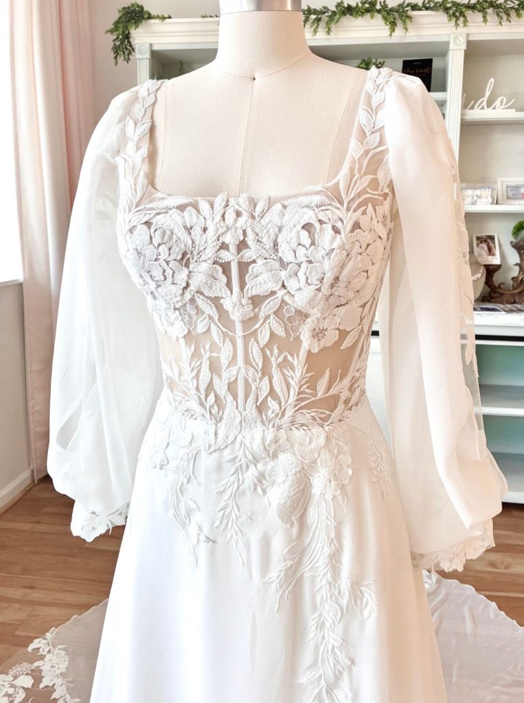 Romantic wedding gown with lace bodice, sheer long sleeves, and square neckline on a mannequin.