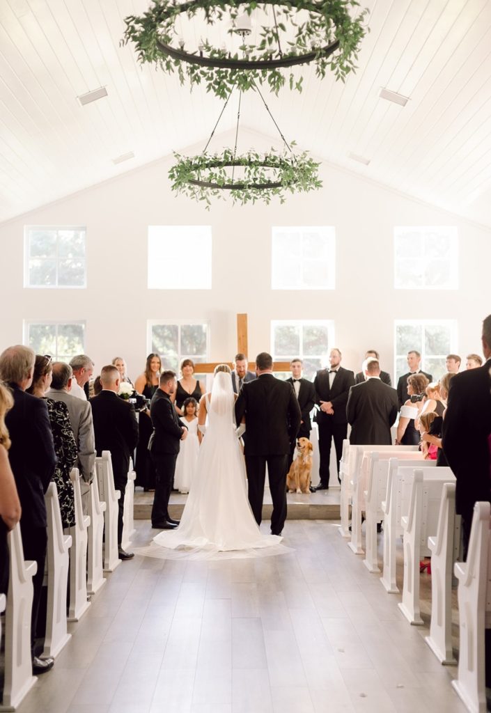 Bride walking down the aisle in a bright chapel filled with guests during a wedding ceremony.