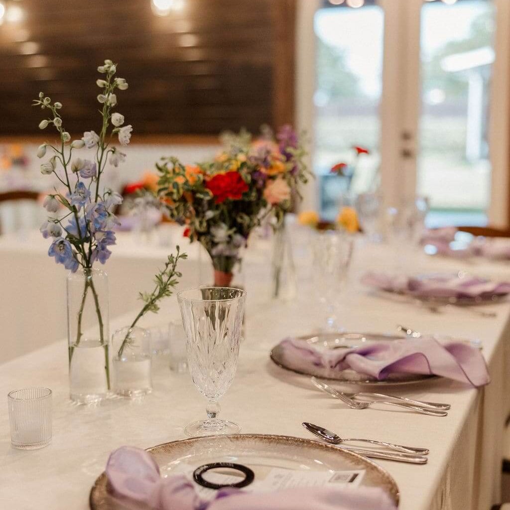 A table setting with flower centerpieces, beige tablecloth, and purple napkins.