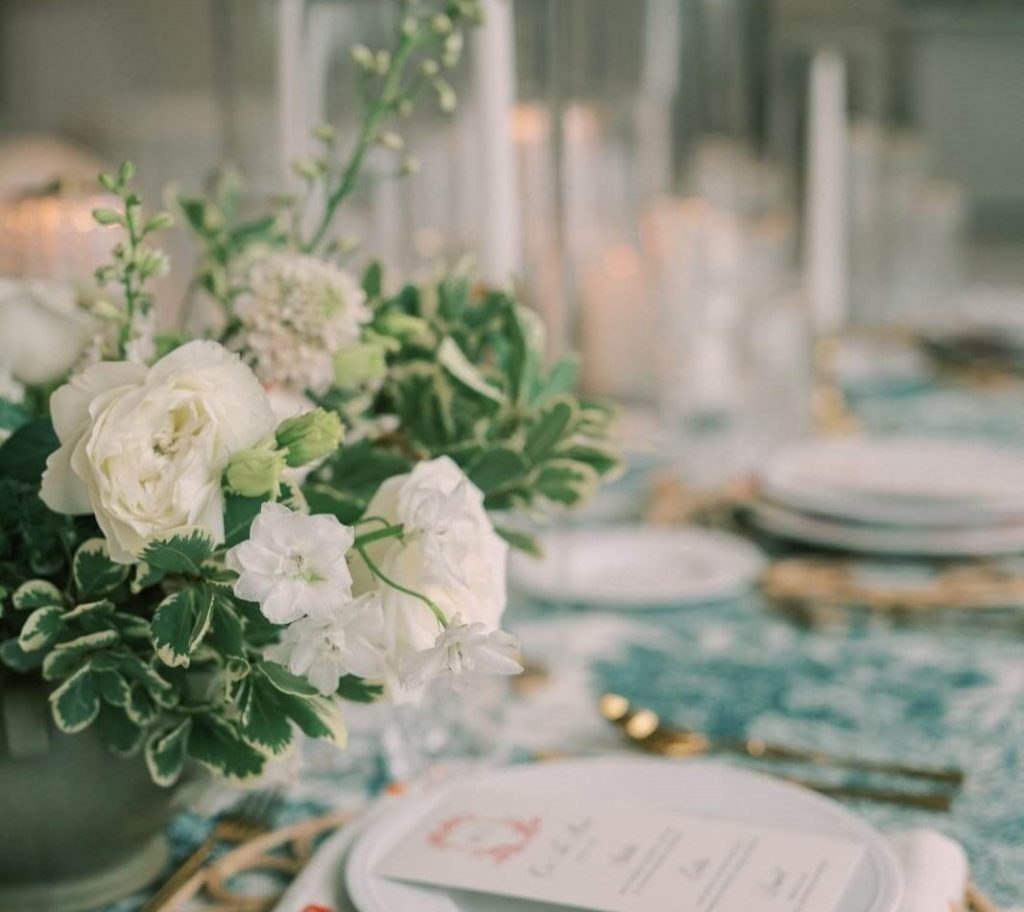 A table setting with white flowers as a centerpiece, white plates, gold cutlery, and a wooden plate mat.