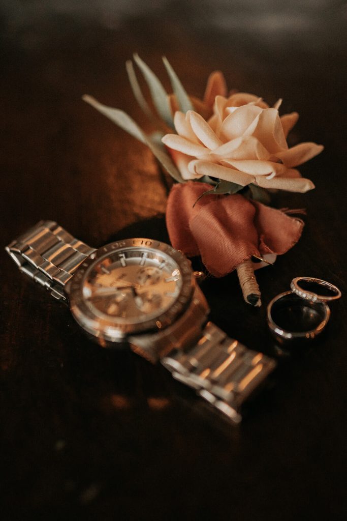 A silver watch, two wedding bands, and a boutonnière lay on a dark wood table.