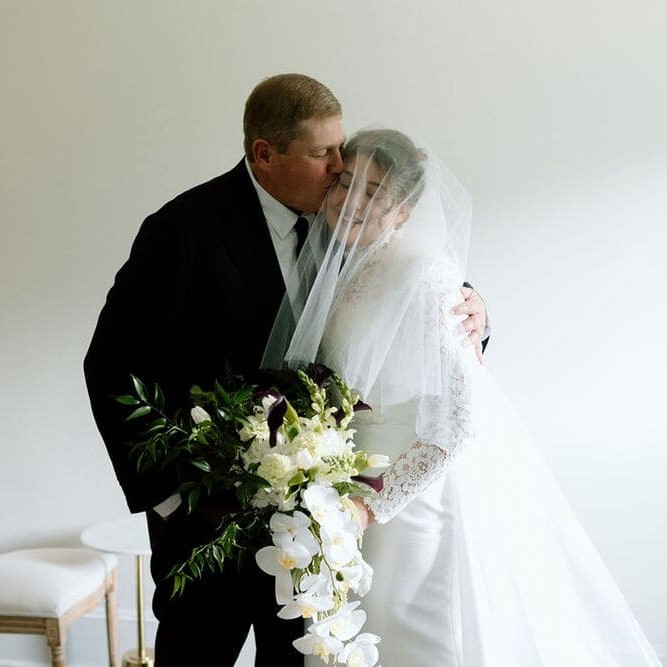 A bride dressed in her wedding gown and veil is holding a bouquet of white flowers while her father gently kisses her on the cheek.