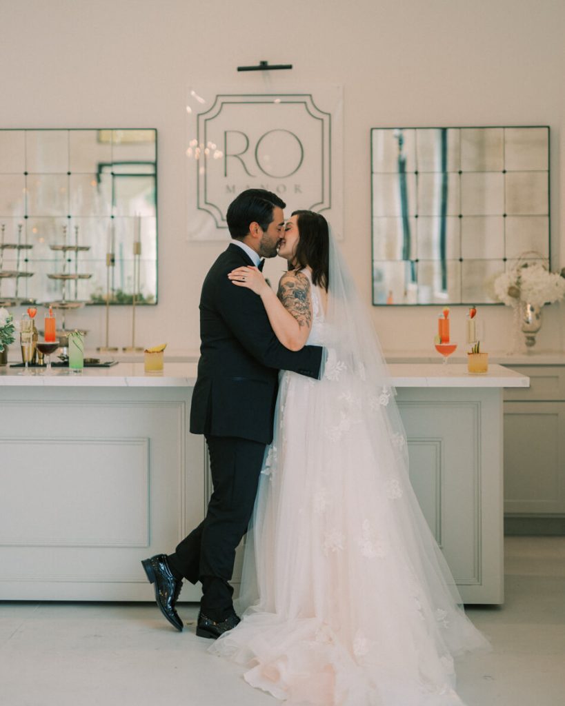 A groom and bride kissing by the cocktail booth at their wedding venue.