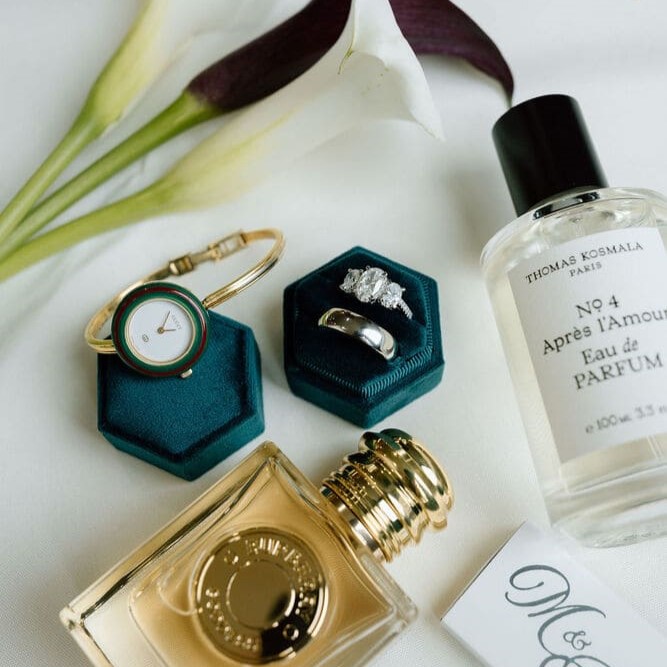 Flat lay of wedding accessories including two rings in teal velvet boxes, a gold bracelet watch, two perfume bottles, calla lilies, and a monogrammed card with initials M and A.