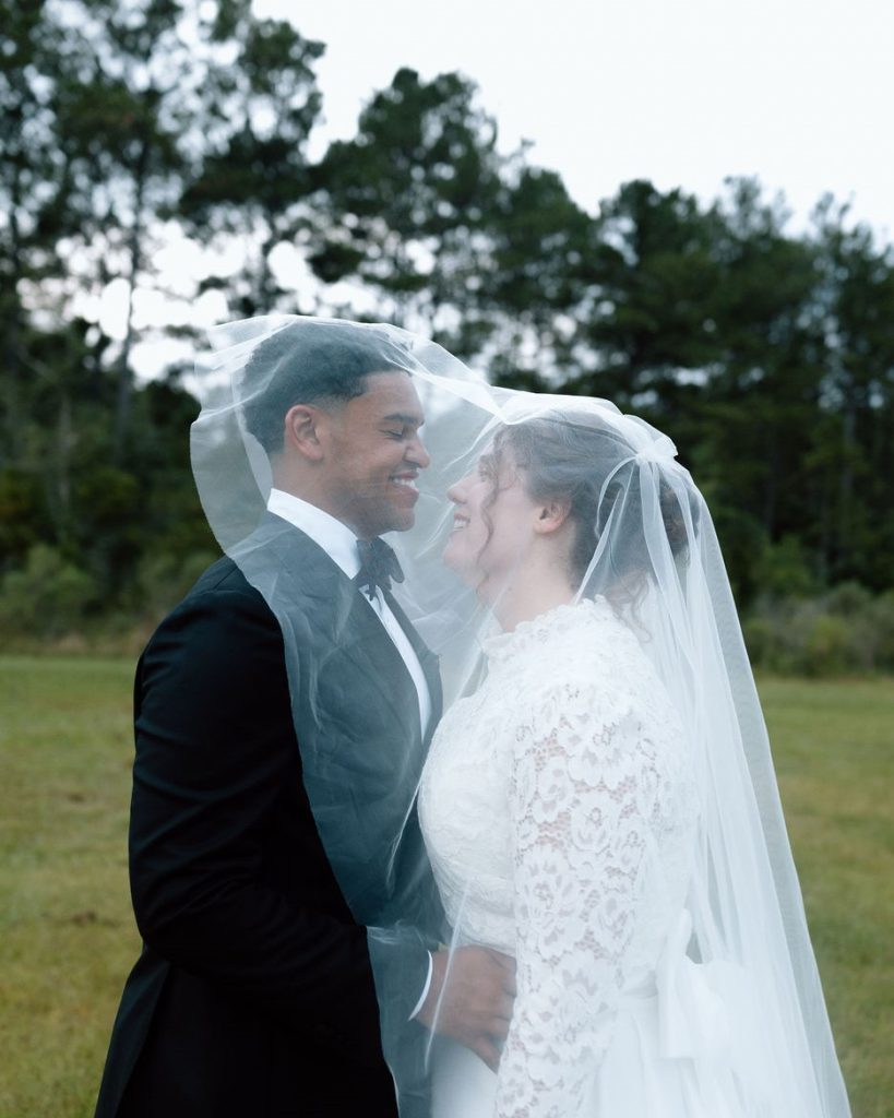 Bride and groom smiling under the bride&rsquo;s veil outdoors, with the groom in a black tuxedo and the bride in a lace wedding dress, standing in a grassy field with trees in the background.