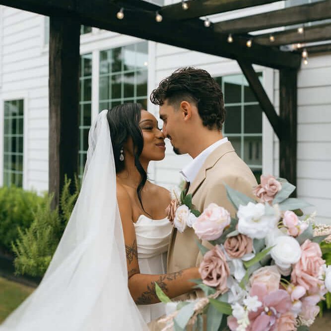 Bride and groom standing close and smiling tenderly under a wooden pergola, the bride in a white gown with a long veil and the groom in a beige suit, holding a bouquet of pink and white flowers.