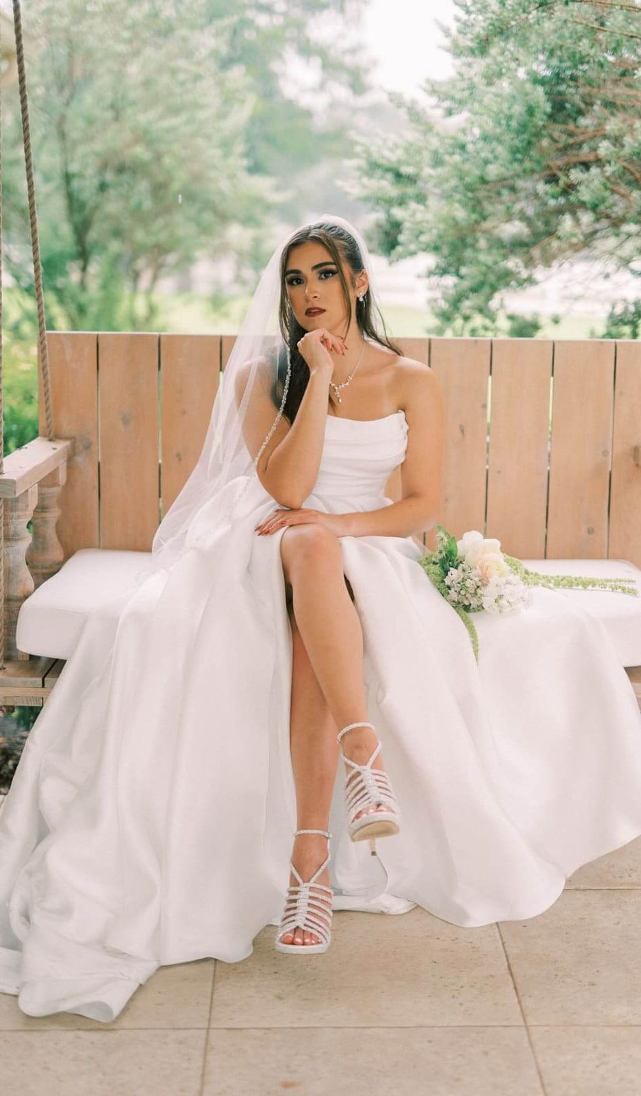 A bride in her flowy white wedding dress is sitting on a wooden swing with her legs crossed and one hand on her chin.