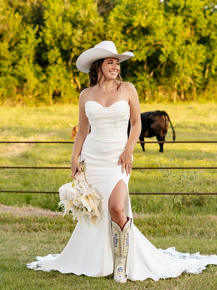 A gorgeous bride standing in the middle of a barn with cows behind her is wearing her wedding dress, cowboy boots, and a cowboy hat, holding a bouquet