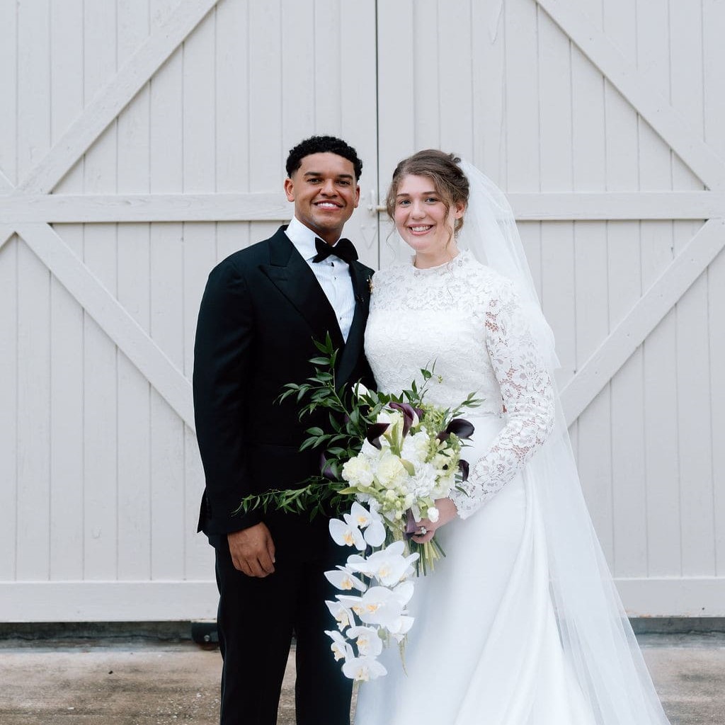 A bride and groom stand smiling in front of a white barn door, holding a large cascading bouquet.