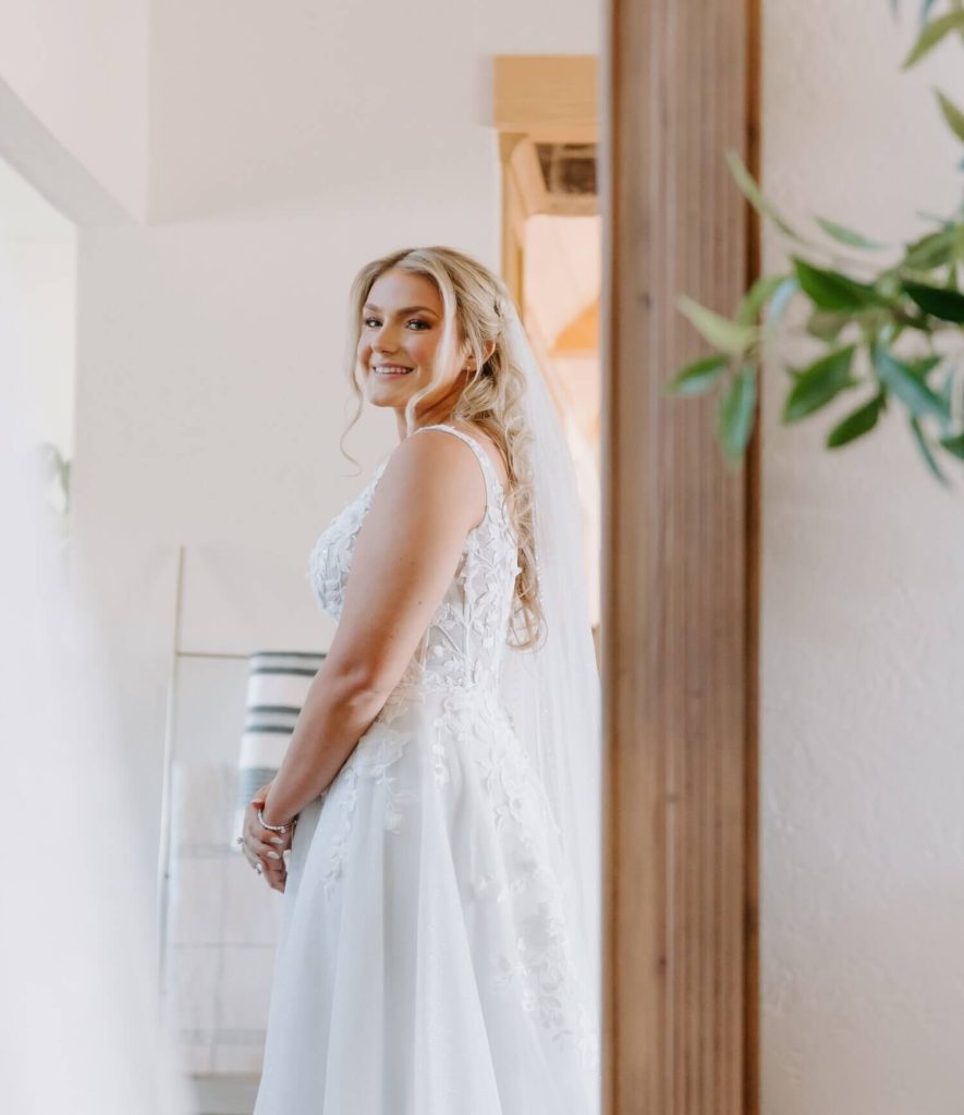 A blonde bride is smiling while looking at herself in the mirror, wearing her A-line wedding dress.