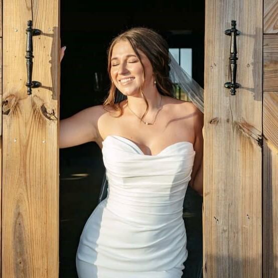 A woman in a fitted wedding dress is opening a wooden door with her eyes closed, shielding her face from the bright light shining toward her.