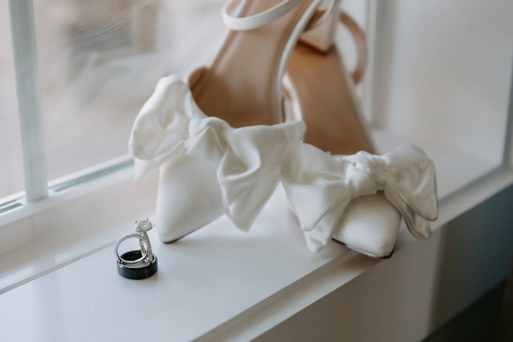 A pair of white satin pointed-toe heels with a bow, accompanied by two rings placed near the window.