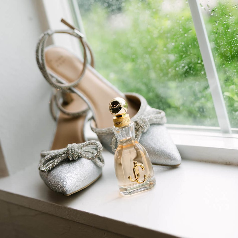 A pair of silver heels and a bottle of Jimmy Choo perfume are positioned near the window, with raindrops visible on the glass.